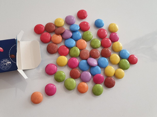 marties box, with the contents of the box spilled out onto the table. There are 8 blue Smarties, 7 yellow Smarties, 8 brown Smarties, 8 red Smarties, 4 purple Smarties, 7 green Smarties, 4 orange Smarties and 5 pink Smarties. In total, there are 51 Smarties.
