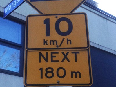 Three street signs on a street. The topmost sign is yellow diamond in shape and shows a speed bump with an arrow above (indicating speed bumps ahead). Below this another yellow sign says 10 km/h in the upper part and Next 180 m on the lower part (indicating the speed bumps continue for the next 180 m of the street and that a driving speed of 10 kilometres per hour is recommended). Below this is a parking sign that says 1P 7.30am-11pm (indicating parking time is limited to 1 hour between those times).
