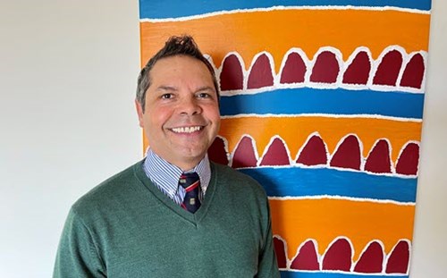 Photo of Professor Chris Lawrence standing in front of a red, blue and orange abstract painting representing mountains and rivers
